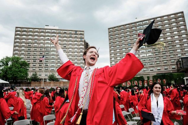 A graduate wearing red robe with colored honor cords holds his hands in the air and cheers.