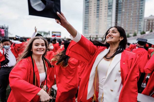 A graduating student wearing a white pantsuit and red graduating gown smiles and holds her cap up in the air mid crowds of graduating students celebrating on Nickerson Field following BU's 2021 Commencement for undergraduate degrees.