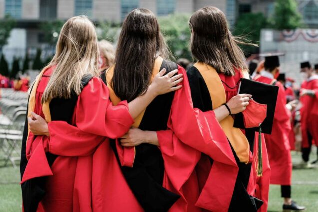 3 women graduates wearing red robes with black and gold stoles photographed from behind with arms around each other's back.