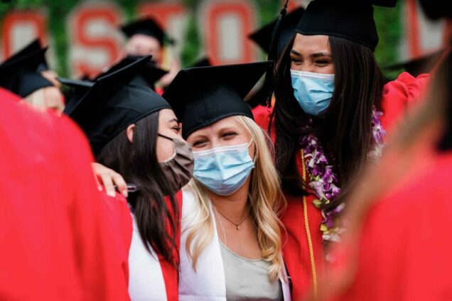 3 graduating seniors wearing graduation caps, gowns and PPE face coverings, lock arms around each other and laugh during BU's 2021 Commencement ceremony for undergraduate degrees.