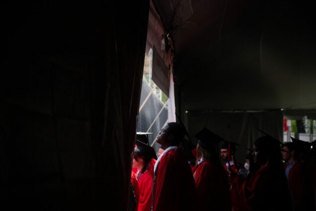 Photo taken underneath the bleachers at Nickerson field. The photo is mostly dark but a door leading out to the field shines bright. A young man of color with a red robe and black mortar is seen processing onto the field at center.
