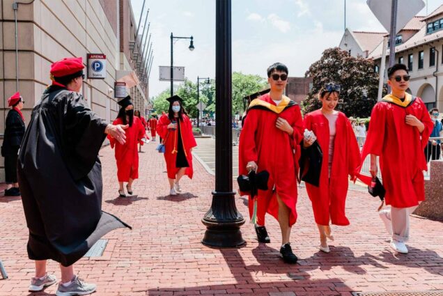 A group of students process down the brick sidewalk along Comm Ave. An AAPI couple, a young man and young woman, are seen at right, another pair of students are seen walking behind them. They all wear the red graduation robes.