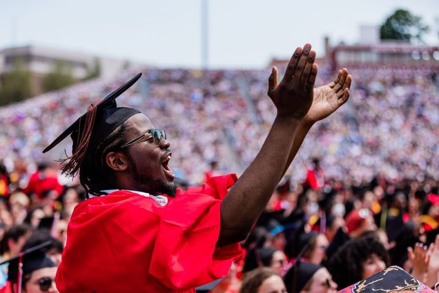 Photo of a young, Black man with a black beard cheering and clapping as he stands amidst a crowd of graduates. He and the grads all wear red gowns and black caps. He also wears stylish sunglasses and a very blurry crowd of people can be seen sitting in bleachers in the background.
