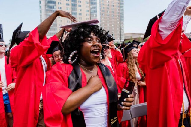 Photo of a young Black person wearing large pearl hoop earrings, a septum nose piercing, and a name necklace that reads "Brittani". hey hold their fist to their chest as they yell with happiness along with the crowd of graduates around them. They all wear red gowns and black caps.
