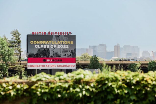 Photo of the Jumbotron at Nickerson field, which reads: Boston University at top. On screen: a photo of Marsh Plaza in Black and White with "Congratulations Class of 2022, BU 2022" overlaid. At bottom, sign reads in red and white: "Congratulations graduates". Hedges are seen in the foreground and the sky is bright blue behind the sign."