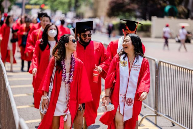 Photo of a group of robbed graduates, two AAPI women and a light-skinned young man with a beard and sunglasses behind them, laugh and talk as they process down Comm Ave. A metal barrier can be seen at left. One woman at right wears a white sash.