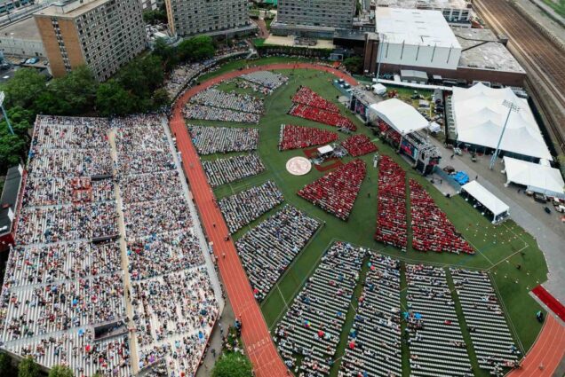 Aerial shot of nickerson field during commencement. Around the stage, at right, is sea of chairs filled with folks in red robes. Behind them, are rows of chairs of attendees and the bleachers, at left, are also filled with people.
