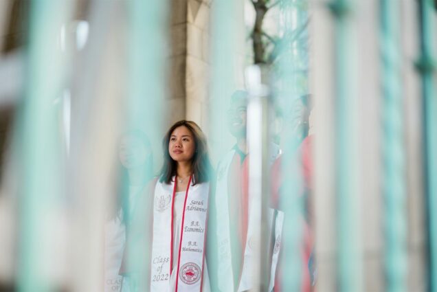 A group of students stand underneath the arches in Marsh plaza. The photo was taken behind white, iridescent streamers so the photo is blurred. A young AAPI woman at center is seen smiling, in focus.