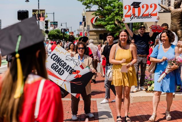 A group of people, possibly a family, stands along the Comm Ave sidewalk holdings that say "Congrats Grad 2022!". At front, are three White women, the middle one wears a yell dress, one at right wears a blue dress. Students processing towards Nickerson can be seen in the foreground and background.