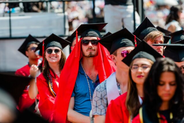 Photo of a light-skinned student at commencement wearing his rob on his head underneath his mortarboard. He wears sunglasses and a blue button down and appears to be wearing his gown on his head to beat the heat. Other students are seen sitting on Nickerson smiling during the ceremony.
