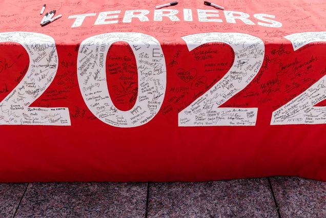 Photo of the red class of 2022 banner, with "Terriers 2022" written in white text, with a bunch of signatures from the class in black. the banner hangs off of a table.