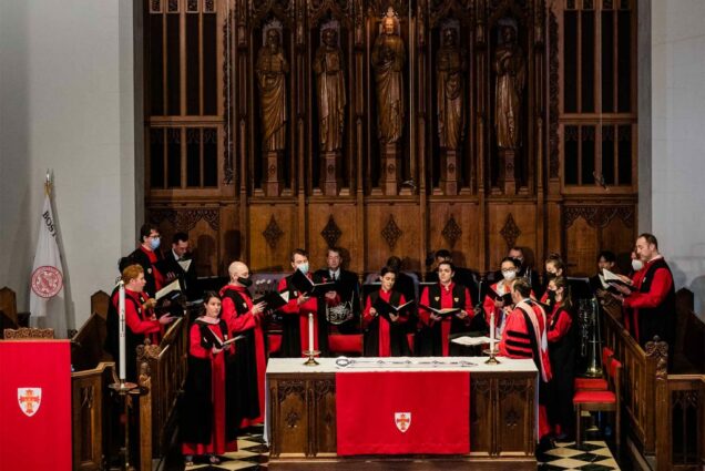 Photo of a group of choir singers dressed in red and black robes during the baccalaureate service. it's a mixture of genders and they all hold open choir books and sign during the service. The large wooden panel is seen behind them.