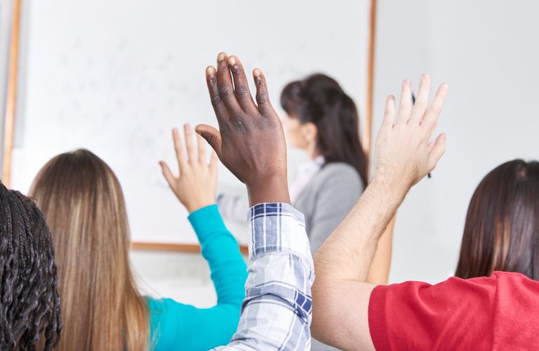 Female professor standing in front of the class, sideways, teaching, and multiple hands raised by students to ask a question