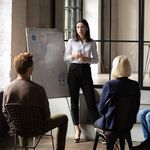 A woman stands by a flip chart presenting to a diverse, seated group in a modern office with large windows, conveying focus and collaboration.