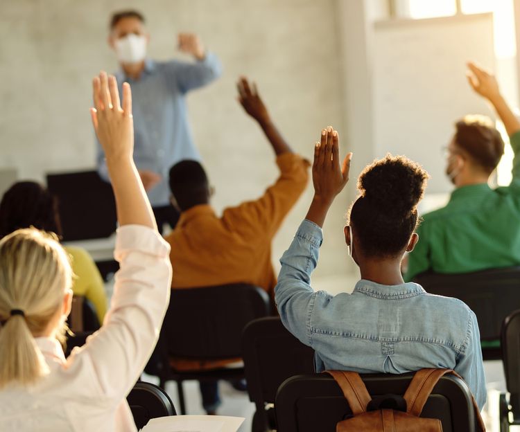 A teacher wearing a mask stands at the front of a classroom with diverse students seated, raising their hands. Sunlight streams in, creating a warm, engaged atmosphere.