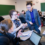 A group engages in a lively classroom discussion. A smiling professor interacts with a student holding a phone, fostering a collaborative mood.