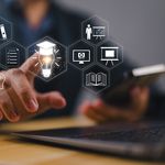 A person is touching digital icons, including a light bulb and target, in front of a laptop, suggesting innovation and technology in a business setting.