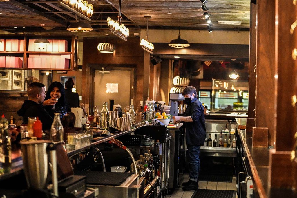 Interior shot of Rosebud Diner. People sit at the wooden bar and a server with a black mask on works behind the counter.