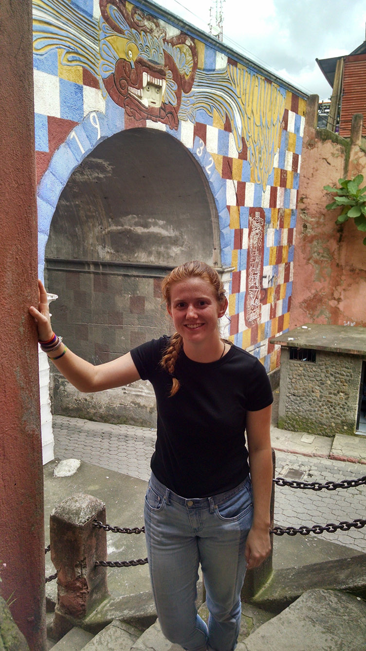 Photo: Student researcher Elizabeth Hannigan, a white woman with long red hair tied into a braid and wearing a black tee shirt and jeans, poses at an archaeological site.