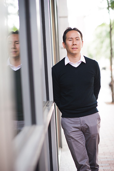 Portrait of Jerry Chen, Boston University College of Arts and Sciences assistant professor of biology, leaning against windows outside a building so that his reflection can be seen to the left.