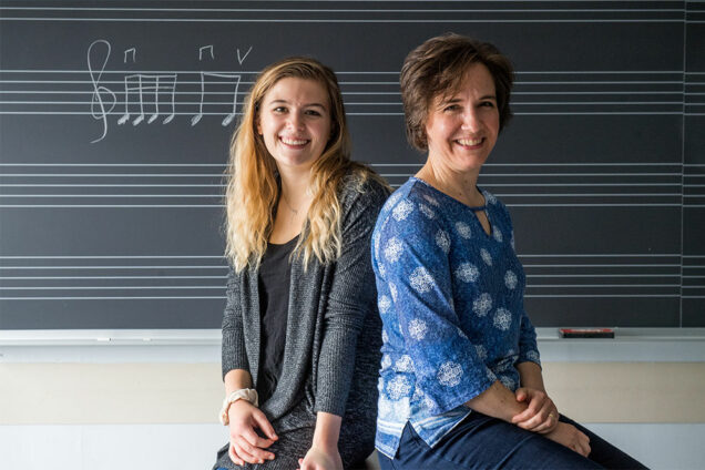 Student Madeline Bucci and College of Fine Arts professor Karin Hendricks sit back-to-back in front of a blackboard with music notes written in chalk.
