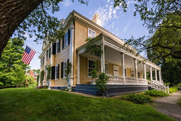 Photo of the Longfellow House Washington’s Headquarters National Historic Site as it looks today. A large burnt yellow house with an American flag posted on its side is shown.