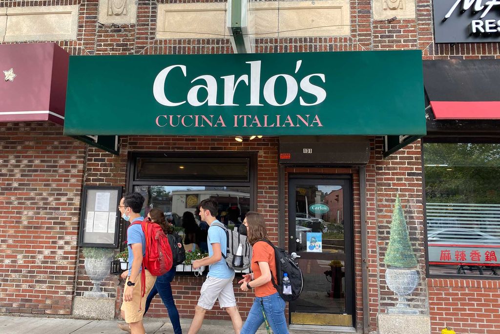 Photo of the exterior of Carlo’s Cucina Italiana, which has a sign with the name on a green awning. People with backpacks on walk by in front of the brick building.