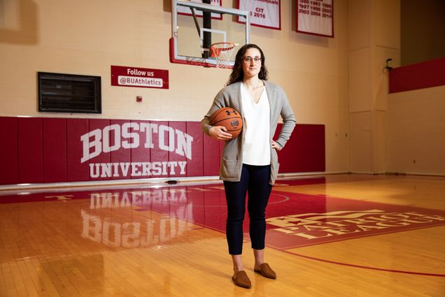 A portrait of Sarah Hope (COM’17) standing with a basketball in front of signs reading "Boston University" and "Follow Us @BUAthletics."