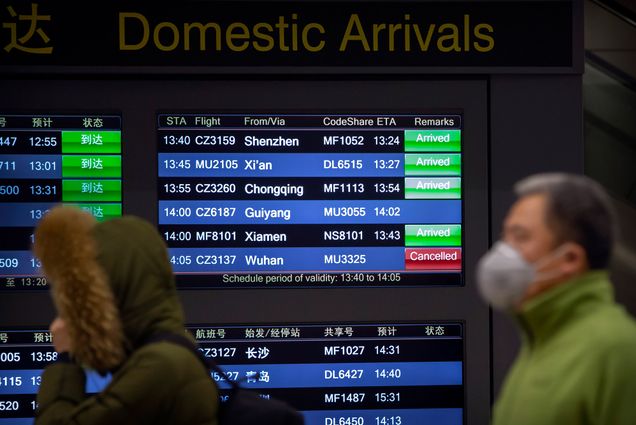 Travelers walk past a display board of arriving flights showing a canceled flight from Wuhan at Beijing Capital International Airport in Beijing.