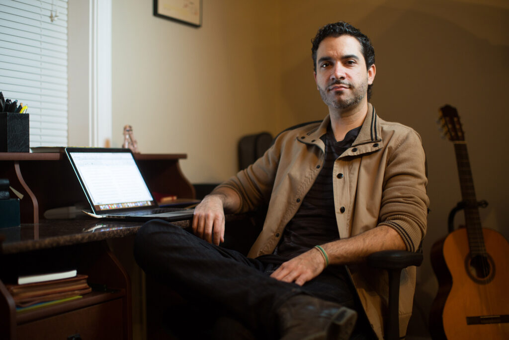 Writer and novelist Dariel Suarez (CFA’12) sits in his office with his laptop open on a desk behind him.