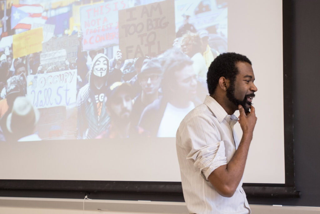 Christopher Rhodes with his hand on his chin while teaching his Identity Politics class. Projector screen behind him shows images of protestors in guy Fawkes masks.