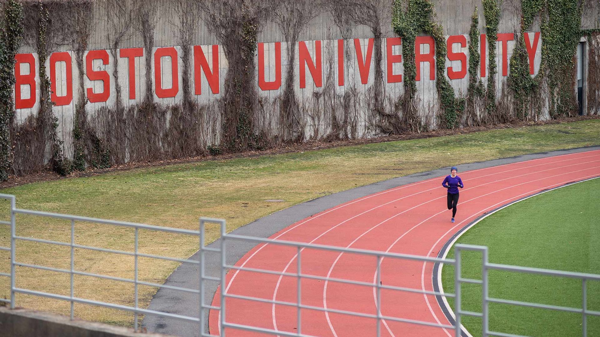 Shot of Nickerson Field with a lone runner in purple jacket running on the track in March of 2020. "Boston University" is seen written on a wall on one side of the track. 