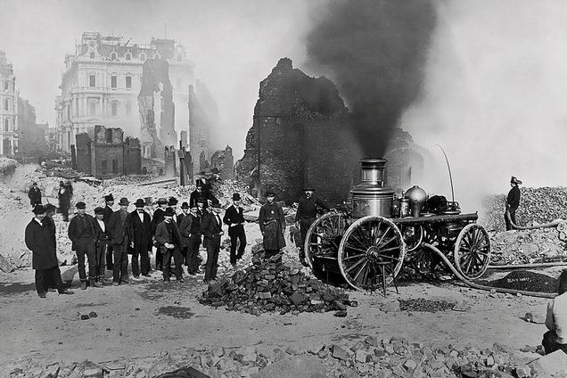 Black and white photograph of the Federal Street Post Office in the wake of the Great Boston Fire of 1872. A billow of smoke rises from the top of an old fire truck, a crowd of men stand close by.