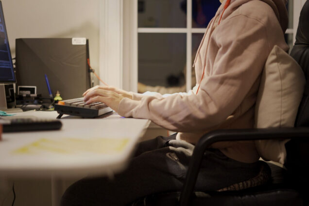 Close-up image of a person working at a home office desk with a pillow behind their back; you can see their hands touching a keyboard.