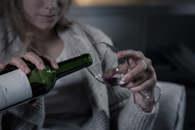 Close-up of a woman's hands as she pours herself a glass of wine; the atmosphere is very dark and gray.