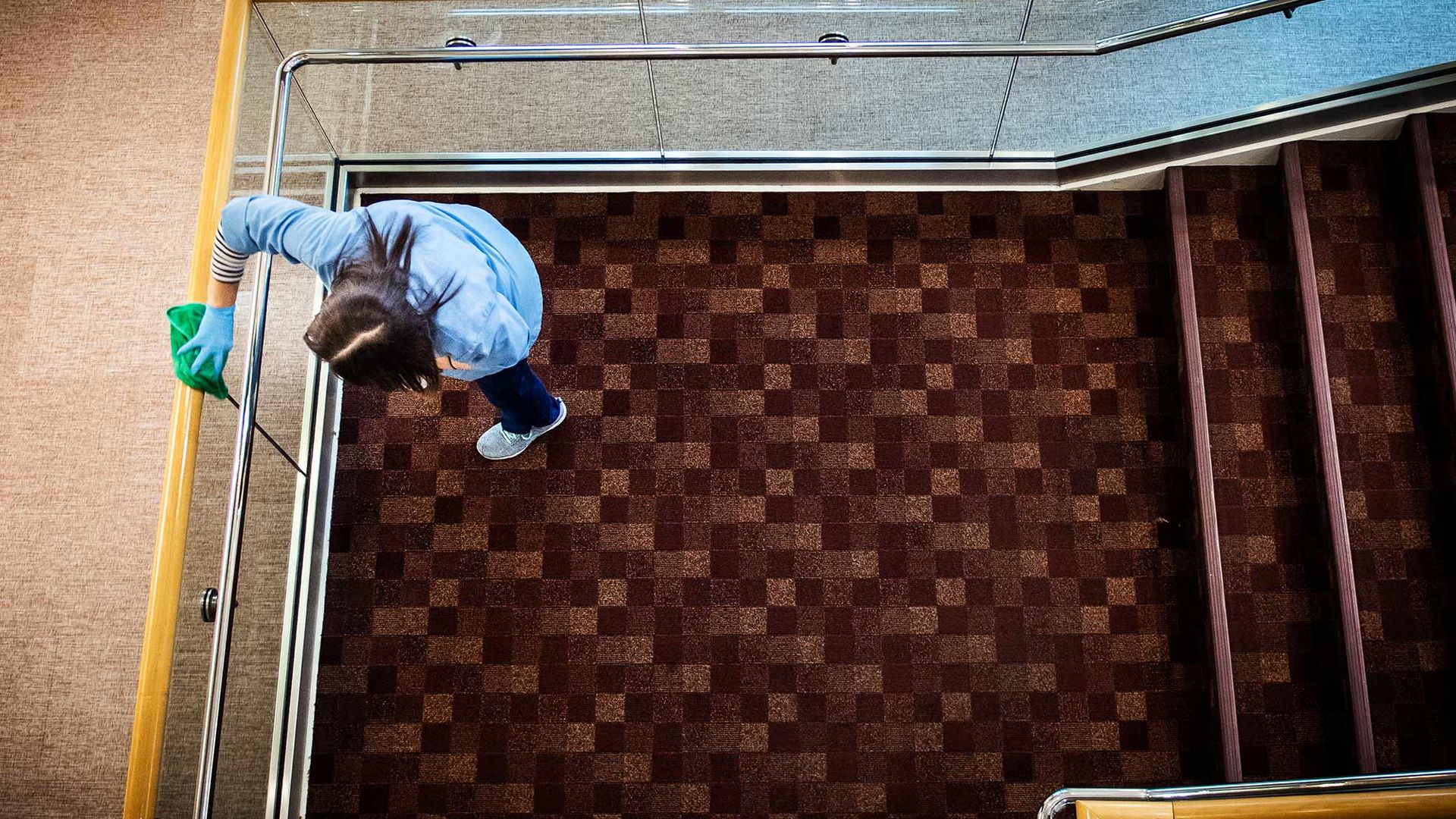 Aerial shot of Grace Araujo of BU Facilities as she cleans StuVi-1 in March 2020. She wears a blue shirt, gloves, and the maroon pattern of the carpet is seen.