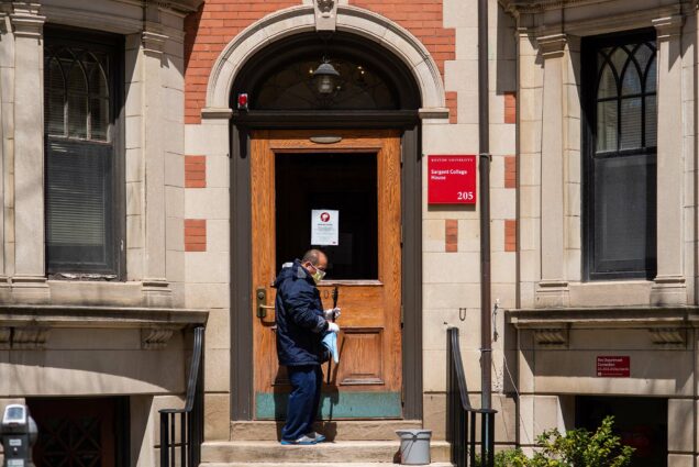 Image of William Lopez, from the facilities department at BU, as he cleans the windows on Bay State Rd on April 29, 2020. A sign on the door reads "important notice" and then red sign on the brick building reads "Boston University Sargent College House 205.