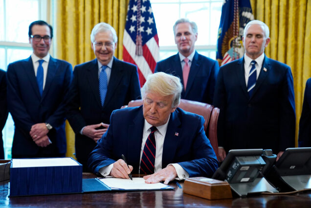 Image of President Donald Trump as he signs the coronavirus stimulus relief package, at the White House, Friday, March 27, 2020, in Washington DC. In the background, from the left, stands Treasury Secretary Steven Mnuchin, Senate Majority Leader Mitch McConnell of Ky., House Minority Kevin McCarthy of Calif., and Vice President Mike Pence, look on. All the men are wearing suits, some are smiling.