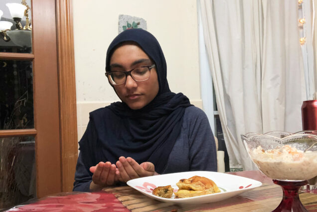 Photo of Azanta Thakur (Sargent’20) prays before breaking her Ramadan fast at her Florida home. A plate of food is before her outstretched hands, and her eyes are closed.