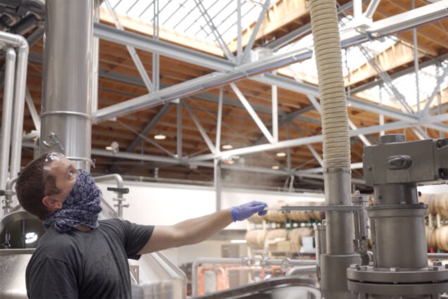 Photo of a brewer from Trademark Brewing producing hand sanitizer during the pandemic. The employee wears a purple face covering and blue gloves and watches a pipe, waiting with his hand on a lever to close the pipe. Barrels are seen in the background.