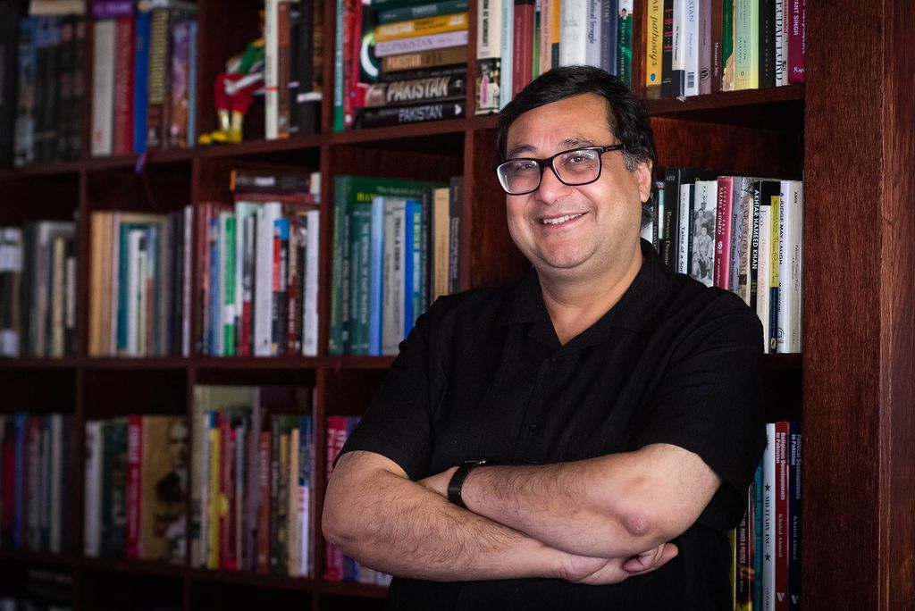 A photo of Adil Najam standing in front of a bookcase in his home with his arms crossed in front of himself