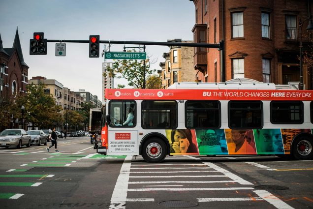 Photo of a BU shuttle driving through an intersection along Mass Ave. The bus has colorful photos on it and a cross walk is seen on the pavement. A pedestrian is seen crossing the street in the distance.