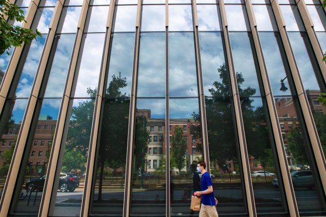 Photo of cloudy blue skies reflect in the windows at CILSE where COVID testing is also being made available to members of the BU community August 19. A student in a mask holds a bagged lunched and walks by.