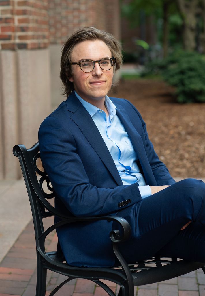 Richard Giadone sits on a bench in a blue suit and smiles. Giadone did his Zoom defense, from his living room in Cambridge, wearing the suit and bowtie his mentor bought him.