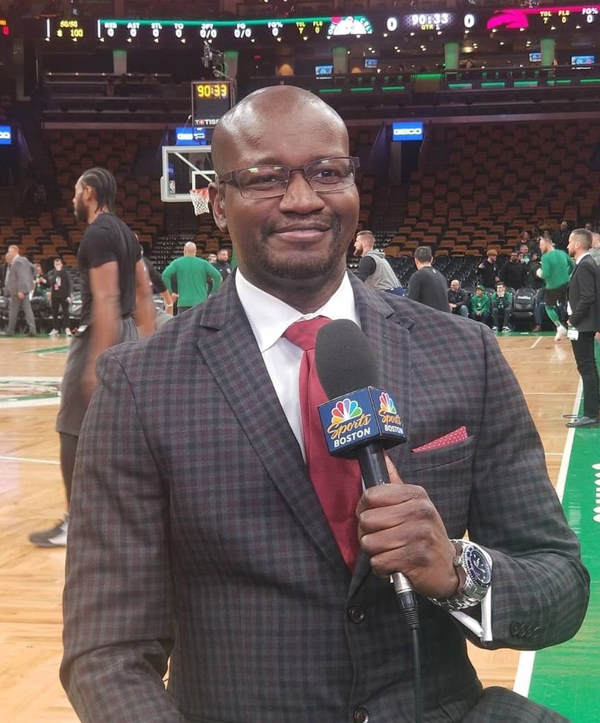 A photo of Sherrod Blakely on the court at TD Garden holding a microphone as NBA players warm up for a game behind him