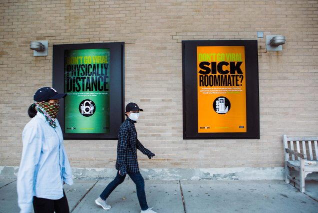 Two students in masks walk by signs on Warren Towers that show ads for BU's 'Don't Go Viral' Campaign. One is green and says “Don’t go viral, physically distance” and another, with an orange background reads “don’t go viral, sick roomate?