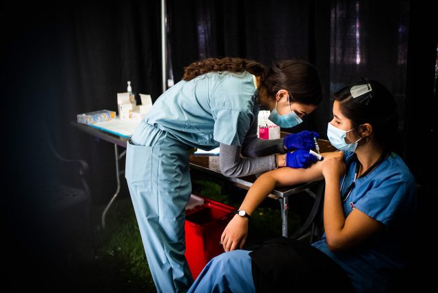 Photo of Renata Mukai (MED’23) getting a flu shot from Sara Khattab (MED’24) during last week’s flu clinic at the Medical Campus. Khattab wears scrubs and both wear blue face masks.