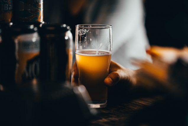 Photo of a hand holding beer; the beer and glass are in the light, while the background is blurry but resembles a bar.