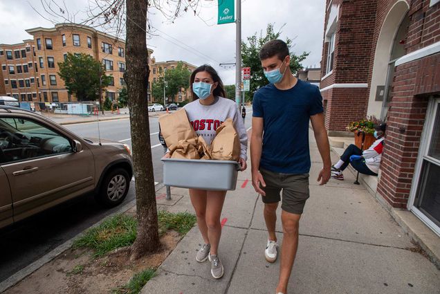 Photo of Flagg (left) and her boyfriend, Aaron Bourget, picking up a donation from Bagelsaurus in Cambridge during a volunteer shift for Rescuing Leftover Cuisine in August. Both wear masks, Lizy carries a tub with brown bags.