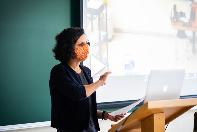 Photo of Caterina Scaramelli teaching an anthropology class, "Culture & Environment “ on September 22, 2020. She wears a bright orange mask as she lectures to her computer and the students in the class. Overlay has video play button.